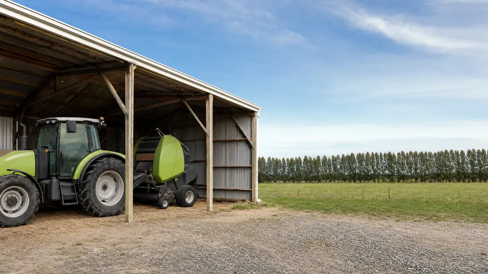 A tractor and round baler parked at the open end of a farm implement shed